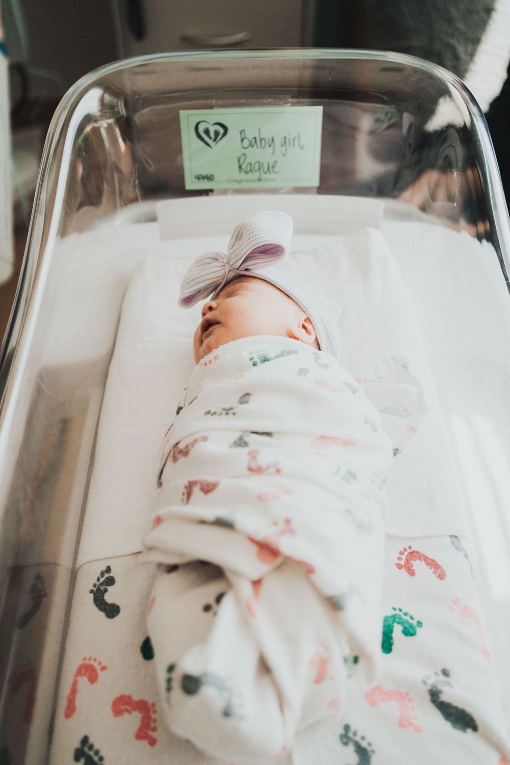 Newborn sleeping peacefully in hospital bassinet wrapped in footprint-patterned swaddle blanket.