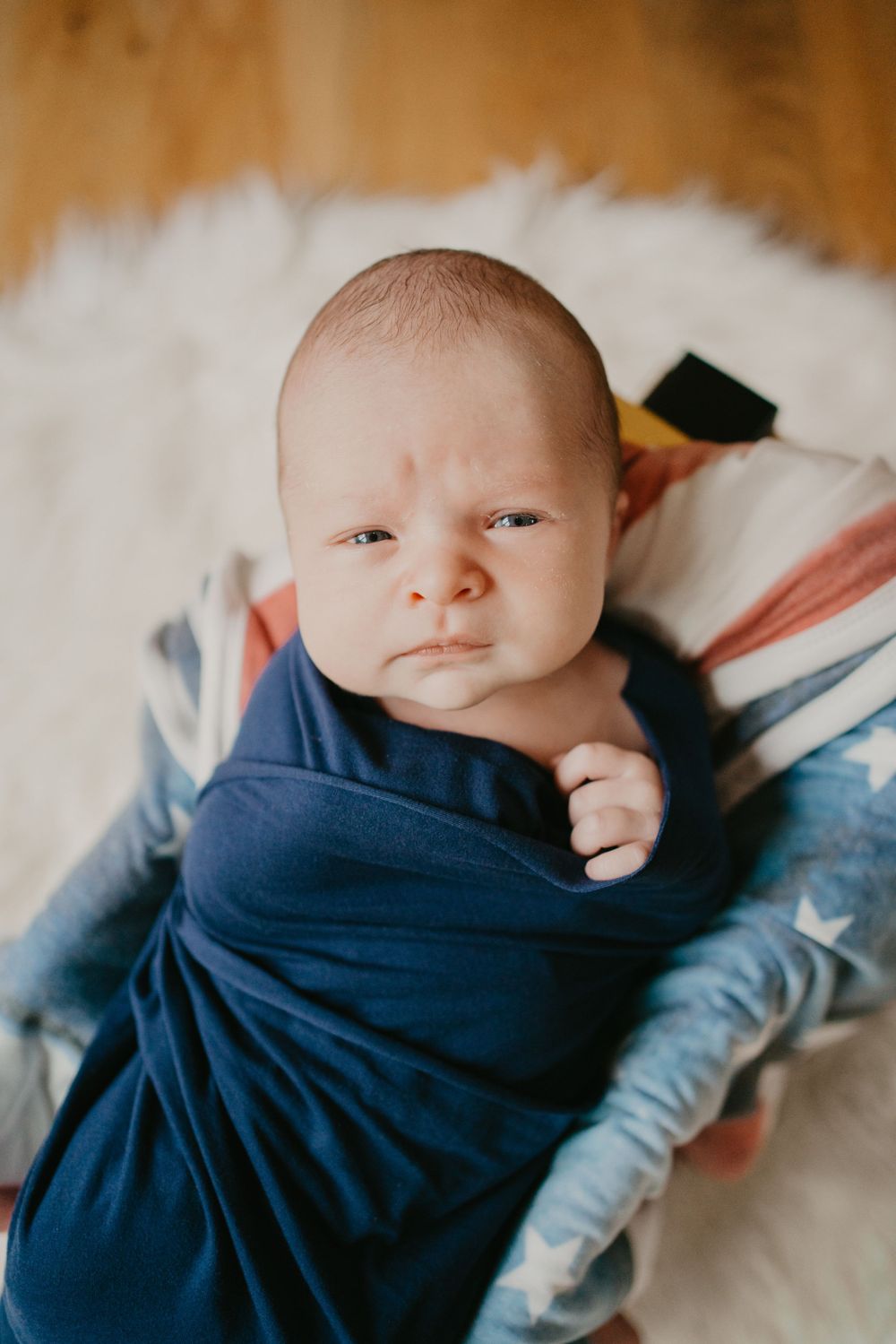 Newborn baby wrapped in navy blue blanket lying on white surface looking directly at camera.