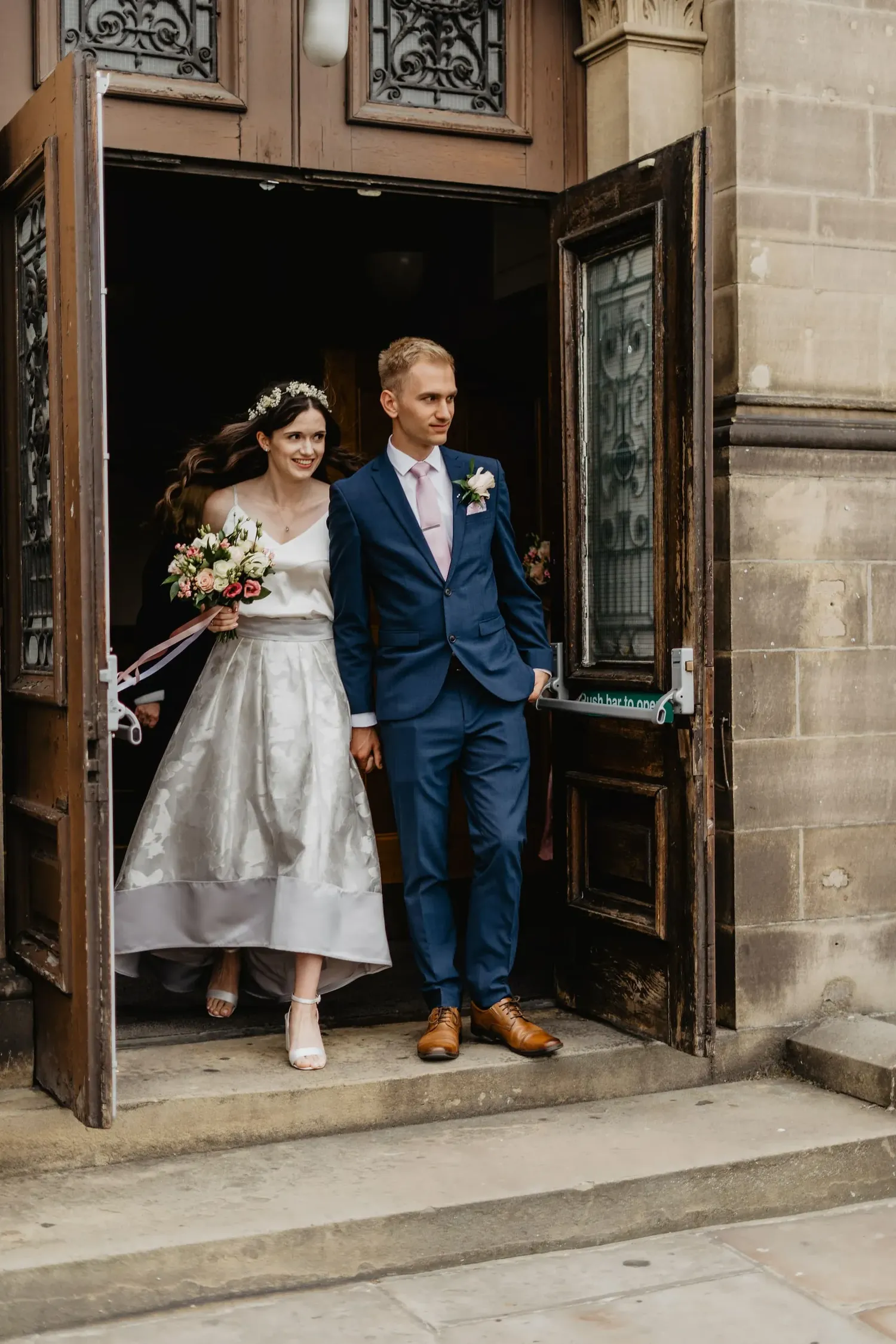 A wedding couple exits an ornate doorway, with the bride in tea-length dress and groom in navy suit.