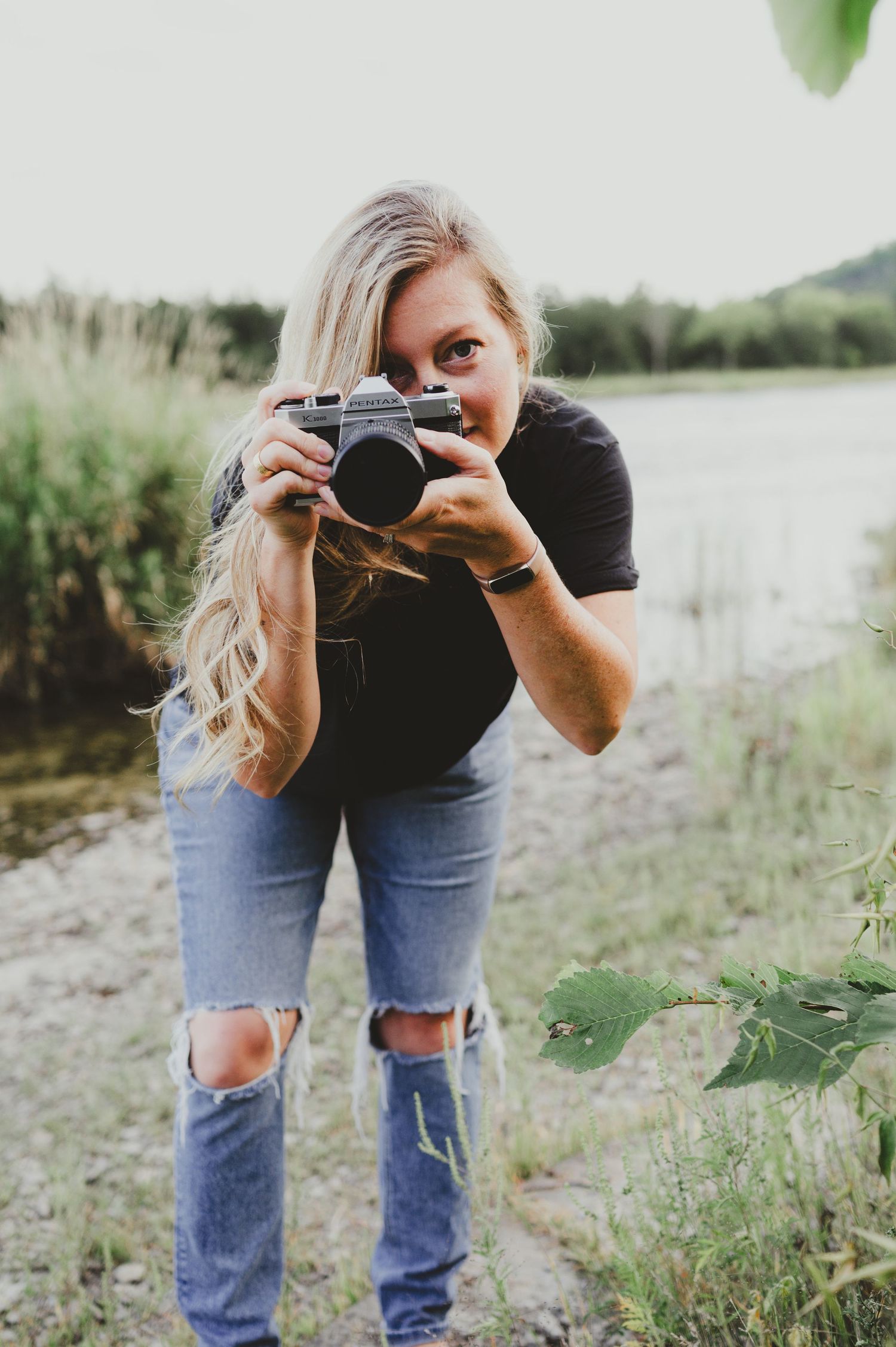 A photographer in ripped jeans takes photos with a vintage camera near a lake.