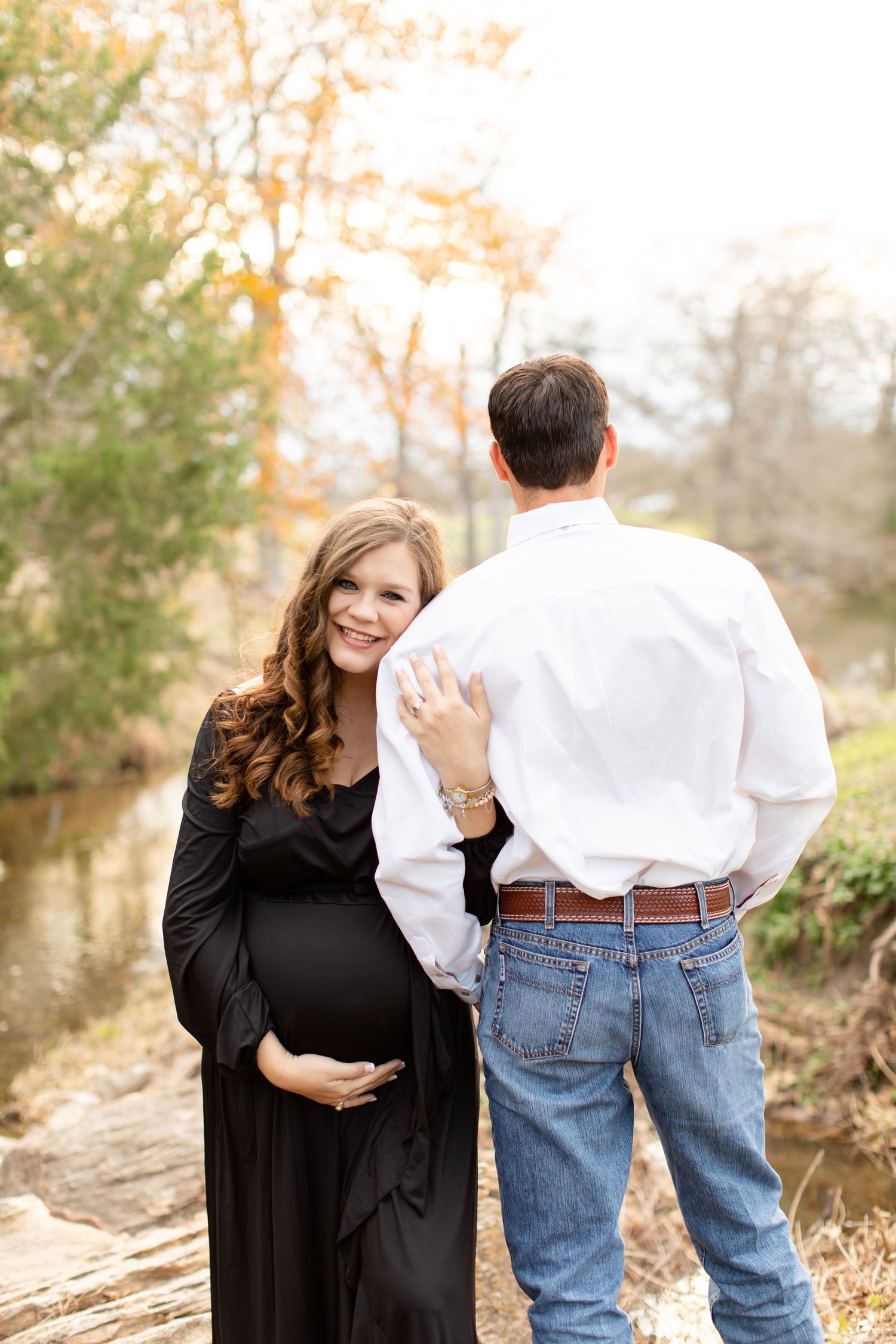 Maternity photo by creek with couple in black dress and white shirt from behind.