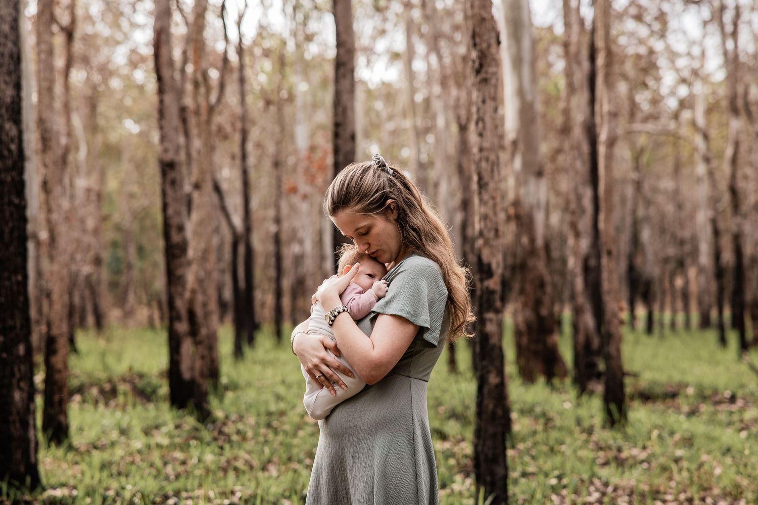 Person cradling an infant in a serene wooded setting with tall trees.