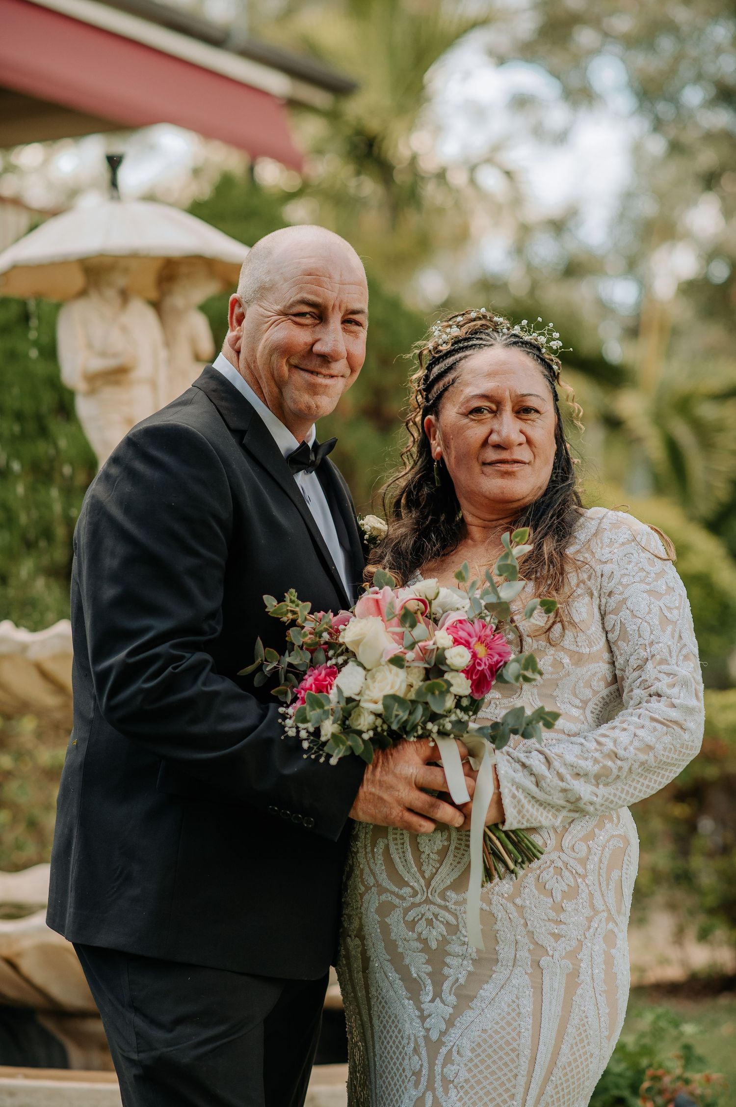 Wedding portrait of couple holding bouquet against garden backdrop with fountain.