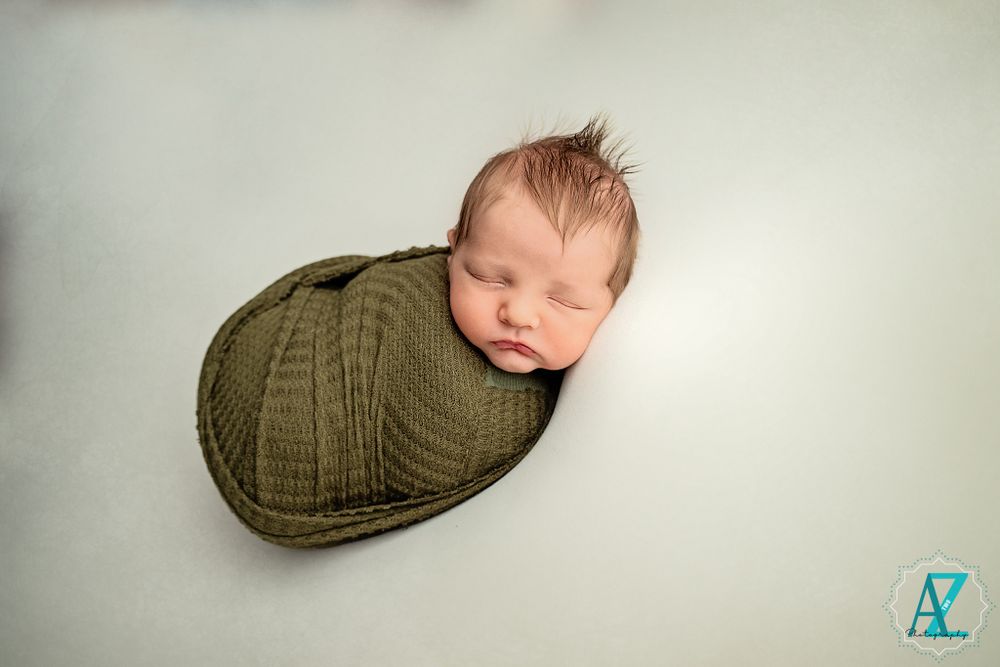 Sleeping newborn wrapped in olive green swaddle on neutral background.