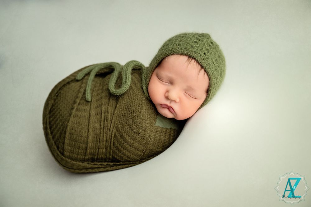 Newborn baby wearing knit green hat and swaddle during professional photography session.