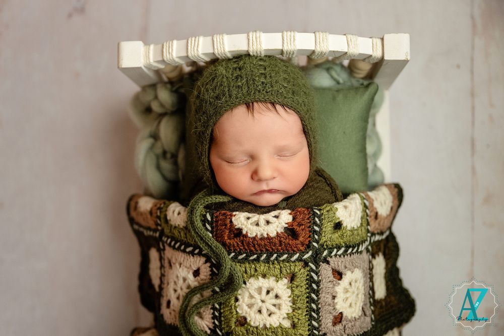 Newborn in green knit clothing sleeps in vintage-style crate with patterned blanket.