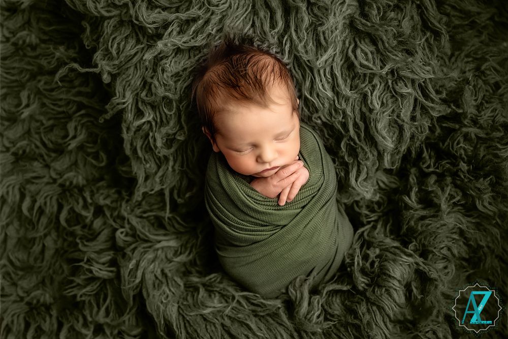 Sleeping newborn swaddled in olive green wrap on fluffy dark green textured blanket.