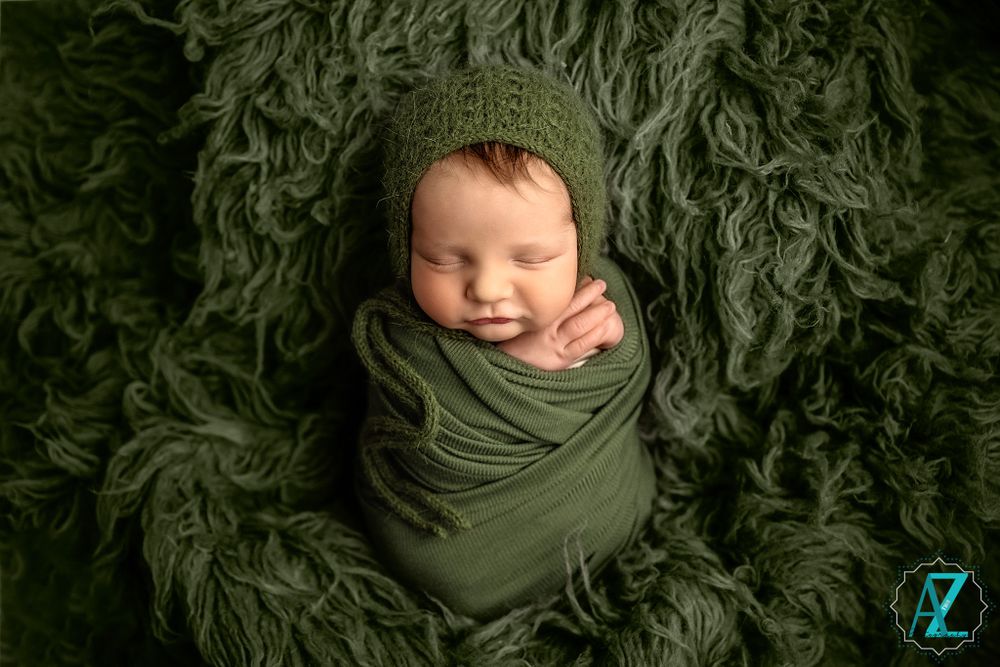 Newborn wrapped in sage green fabric lying on luxurious dark green furry backdrop.