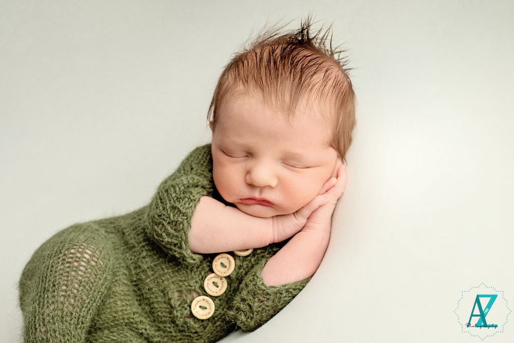 Newborn in olive knit sweater with wooden buttons resting on white backdrop.