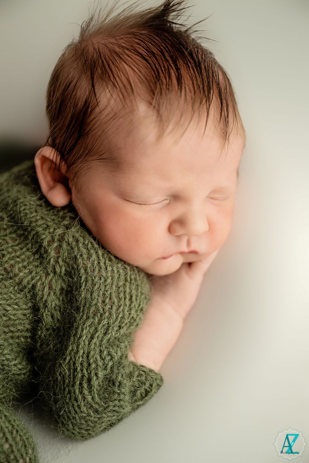 Close-up of sleeping newborn wearing olive green knit sweater on light backdrop.