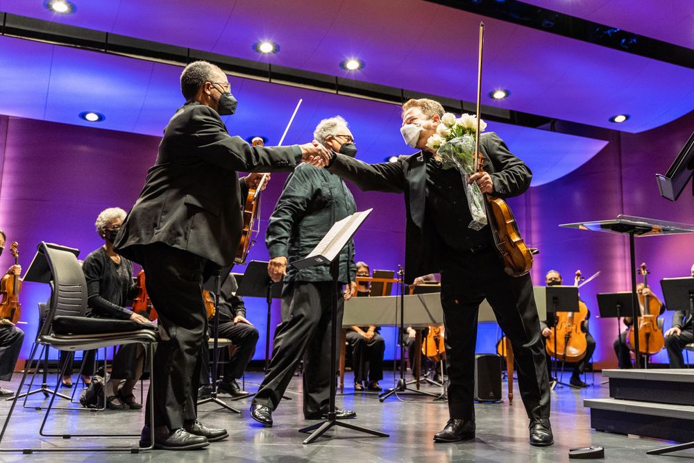 Musicians performing classical music on stage with purple lighting and orchestra in background.