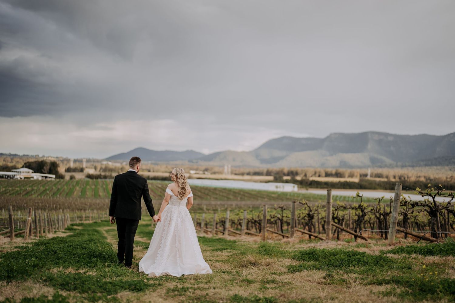 A couple walks through a vineyard with mountains visible in the distance.
