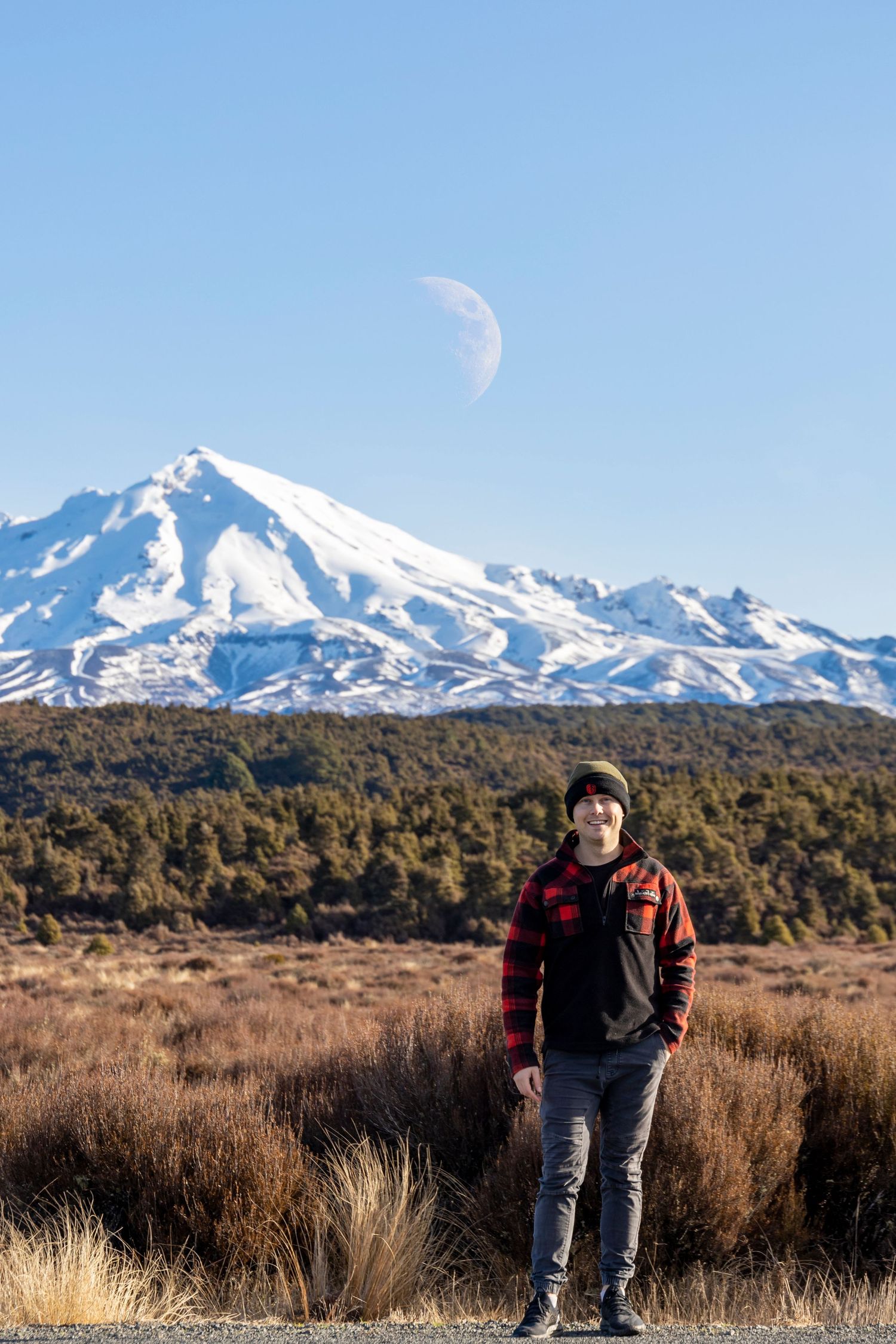 Hiker standing in tussock grass with a snow-capped mountain and crescent moon in the clear blue sky behind.
