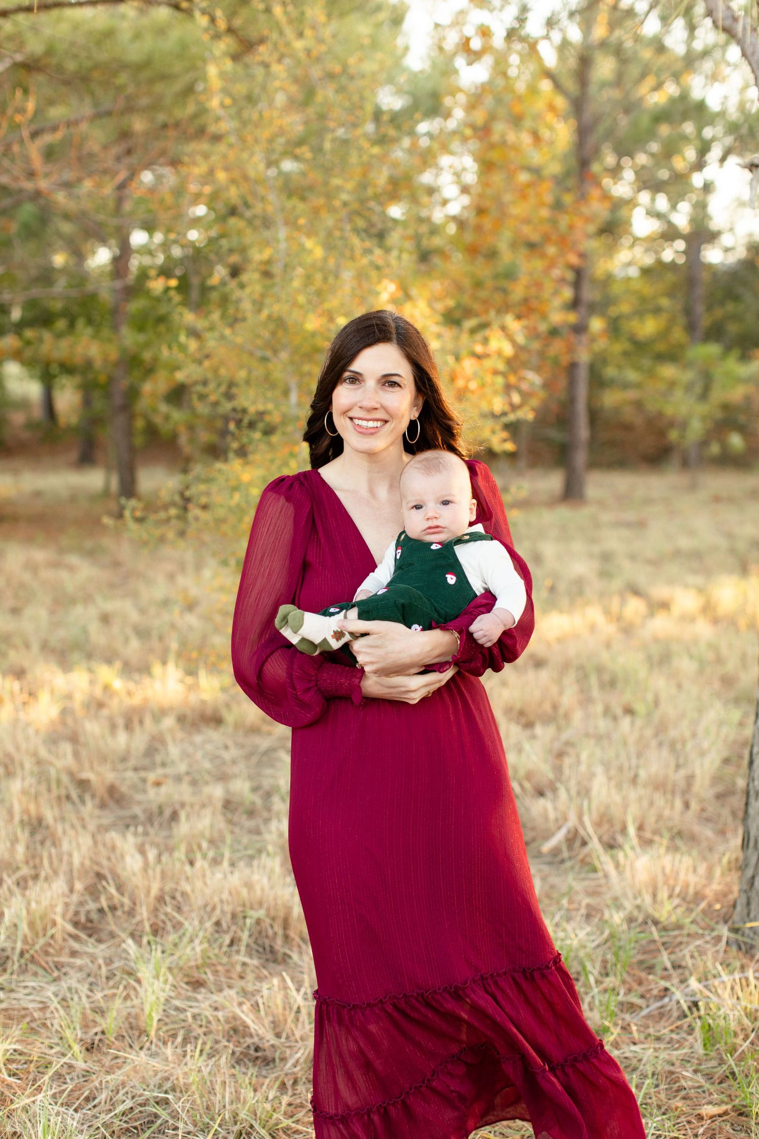 Mother in flowing burgundy dress holding baby in green outfit against autumn foliage.