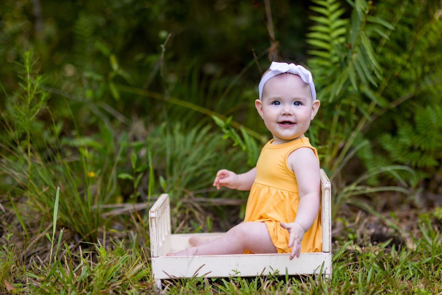 Baby in yellow dress sitting on rustic wooden prop surrounded by ferns.