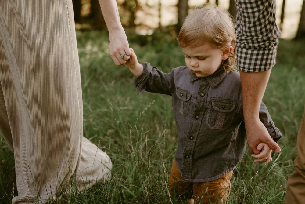 Young child in denim jacket holding hands with adults while walking through tall grass.