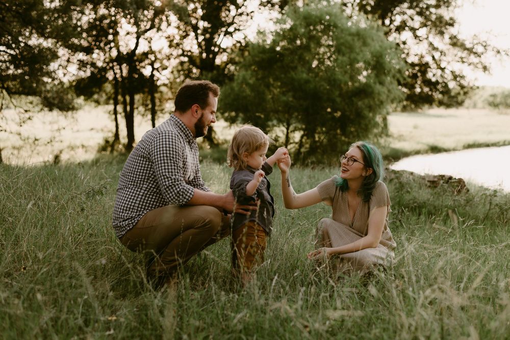 A family shares a sweet moment together in a sunlit grassy field near trees at golden hour.