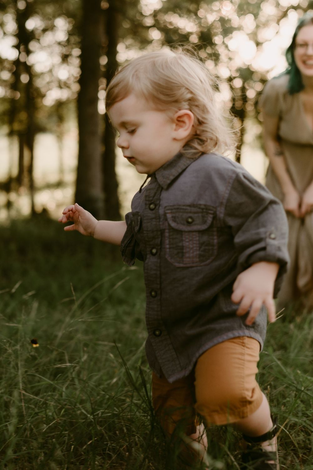 Small child in denim jacket and brown pants exploring outdoors in wooded setting.