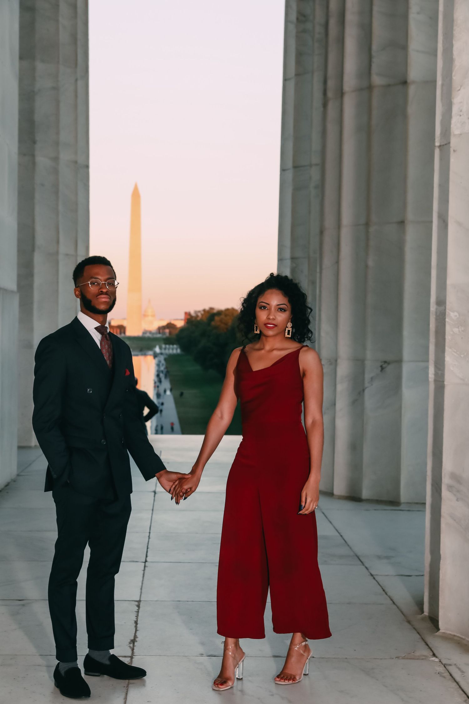 An elegant couple holds hands between marble columns at the Lincoln Memorial with the Washington Monument at sunset.