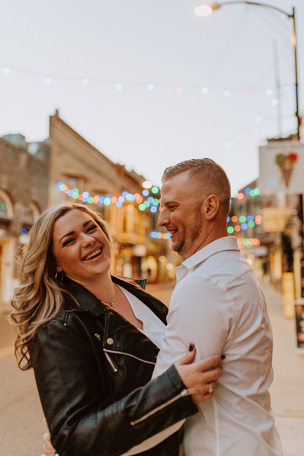 Joyful couple laughing and embracing on small town street with twinkling lights in background.