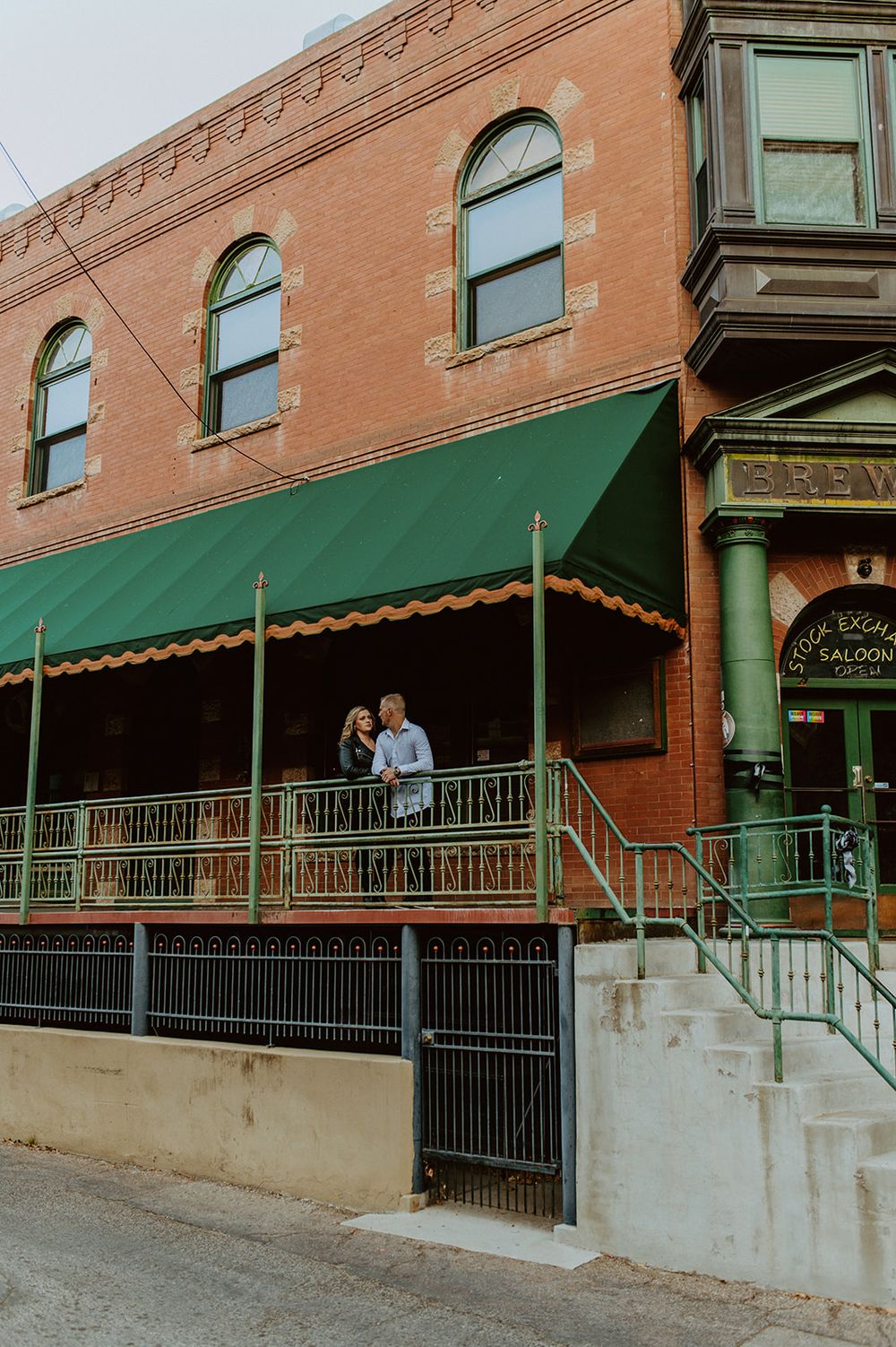 A couple stands on a balcony with green awning and ornate railings of a historic brick building.