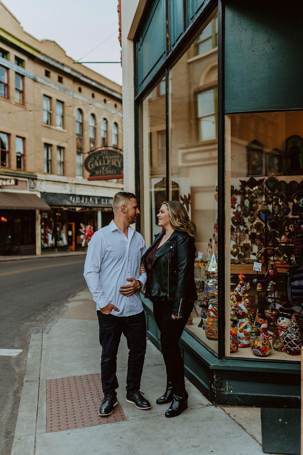 A couple poses together outside a vintage storefront with a display window on a downtown street.