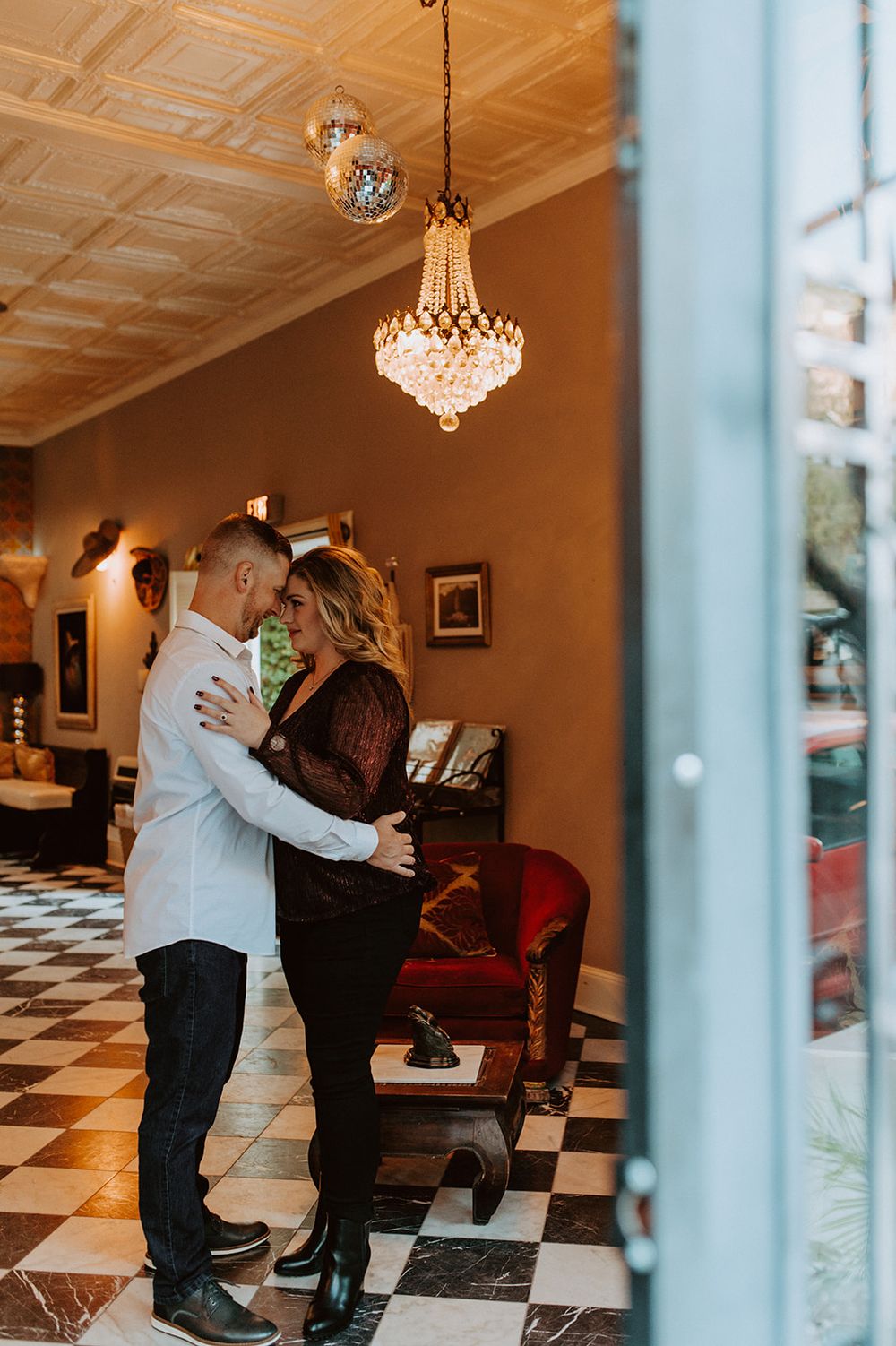 A couple shares an intimate moment under a crystal chandelier in an elegant room with checkered floors.