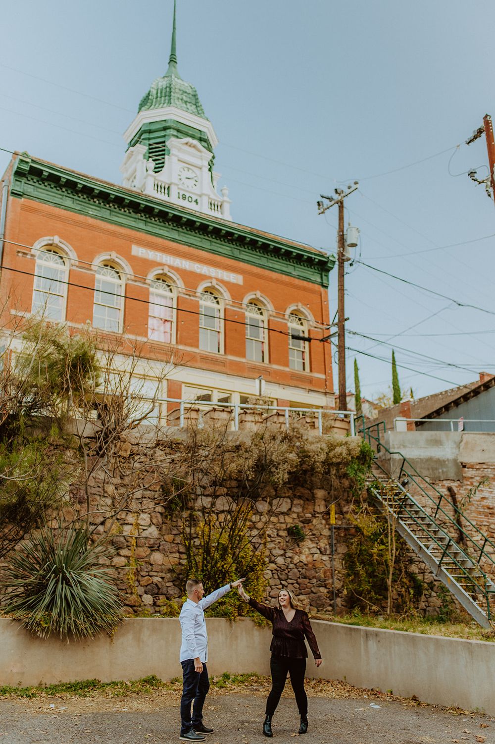 A couple poses playfully in front of a historic orange building with a green spire.
