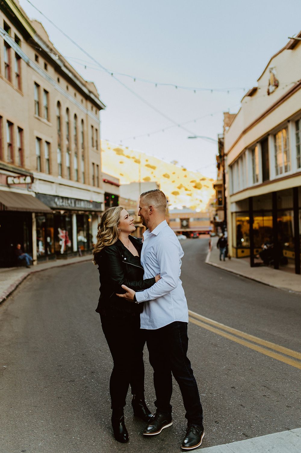 A romantic couple embraces on a city street at sunset with string lights overhead and historic buildings in the background.