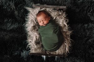 Newborn baby sleeping peacefully on gray fur blanket while wrapped in green swaddle.