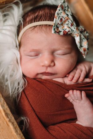 Peaceful newborn wearing floral bow headband resting on rusty red fabric.