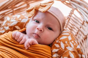 Baby wearing mustard yellow wrap and beige bow headband resting in wicker basket.