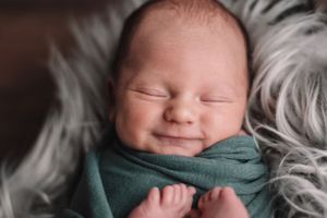 Smiling newborn baby swaddled in green wrap on white furry blanket.