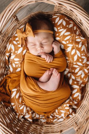 Sleeping baby in mustard wrap with bow headband nestled in wicker basket on floral pattern.