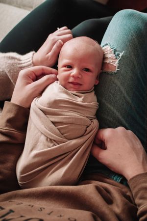 Smiling baby wrapped in beige swaddle lying on caregiver's lap.