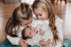 Two children in white sweaters lovingly admiring a swaddled newborn.