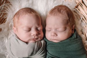 Twin newborns sleeping side by side wrapped in gray and green swaddles on white fur.