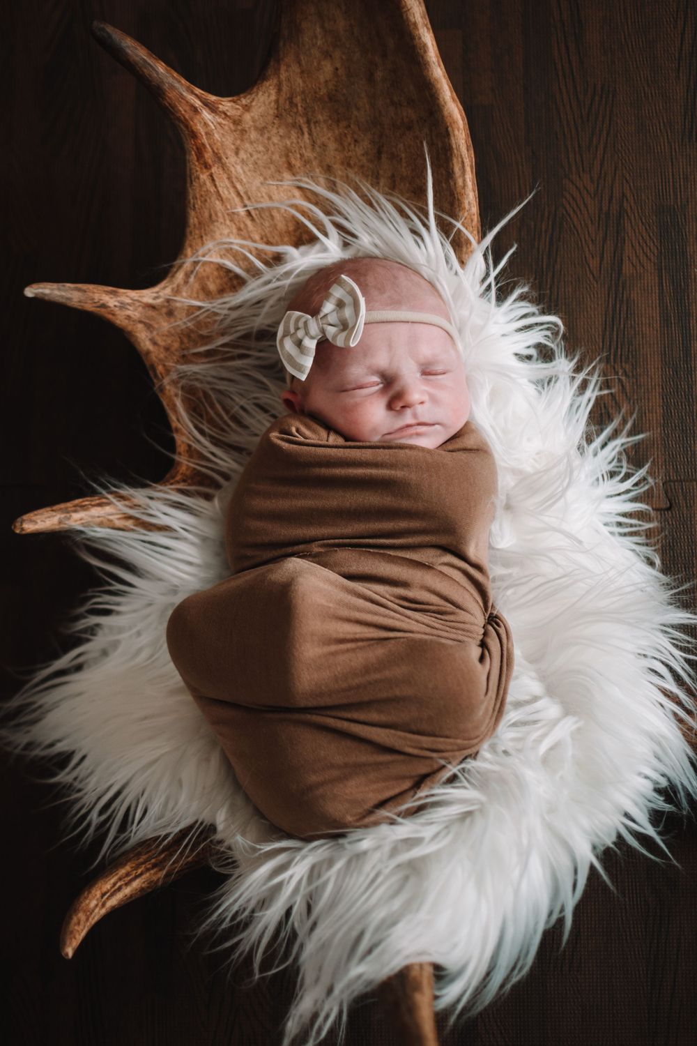 Sleeping newborn wrapped in brown swaddle on white fur with antler decor.