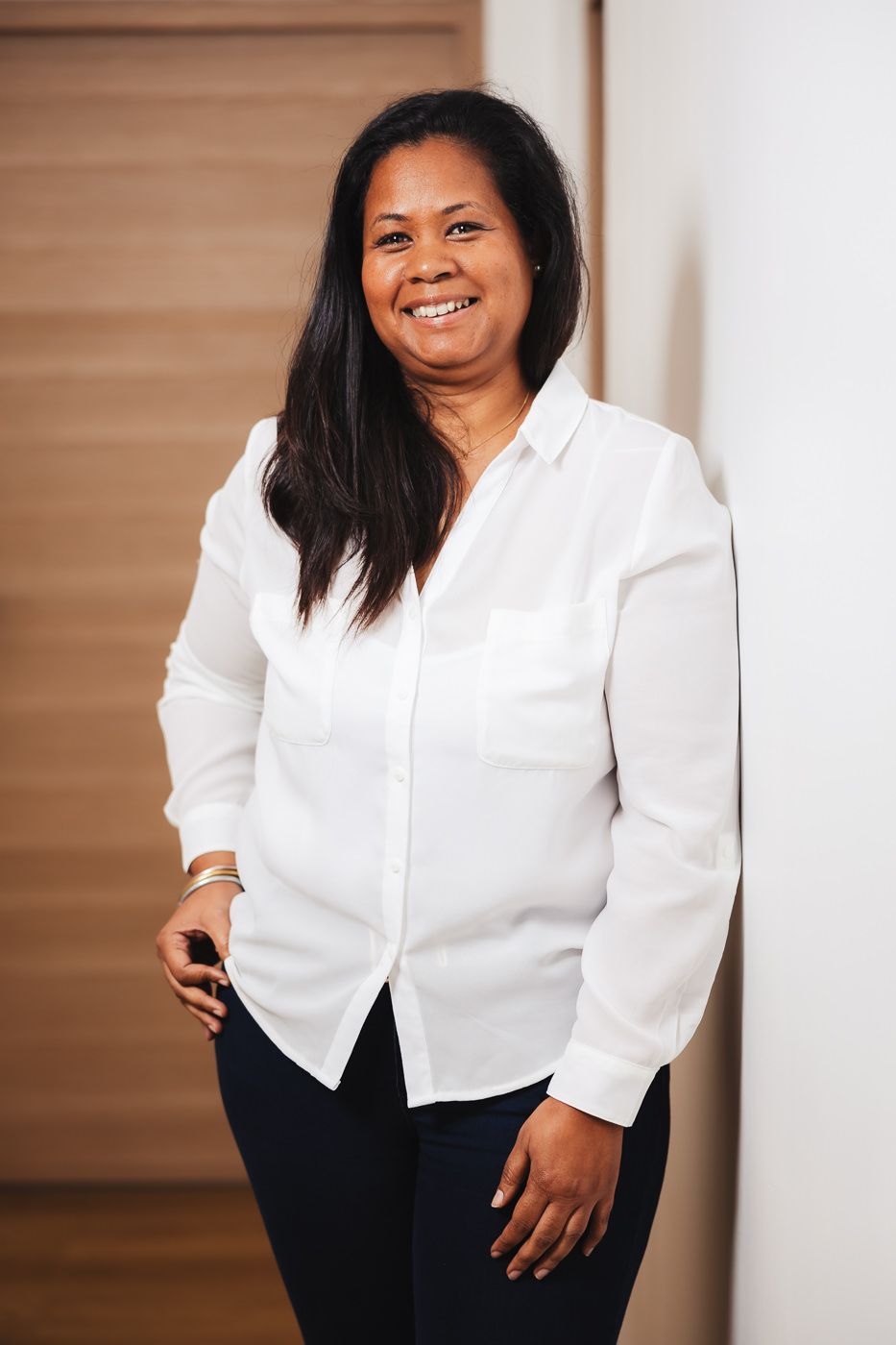 Professional woman in white button-down shirt and black pants smiling warmly against wooden background.