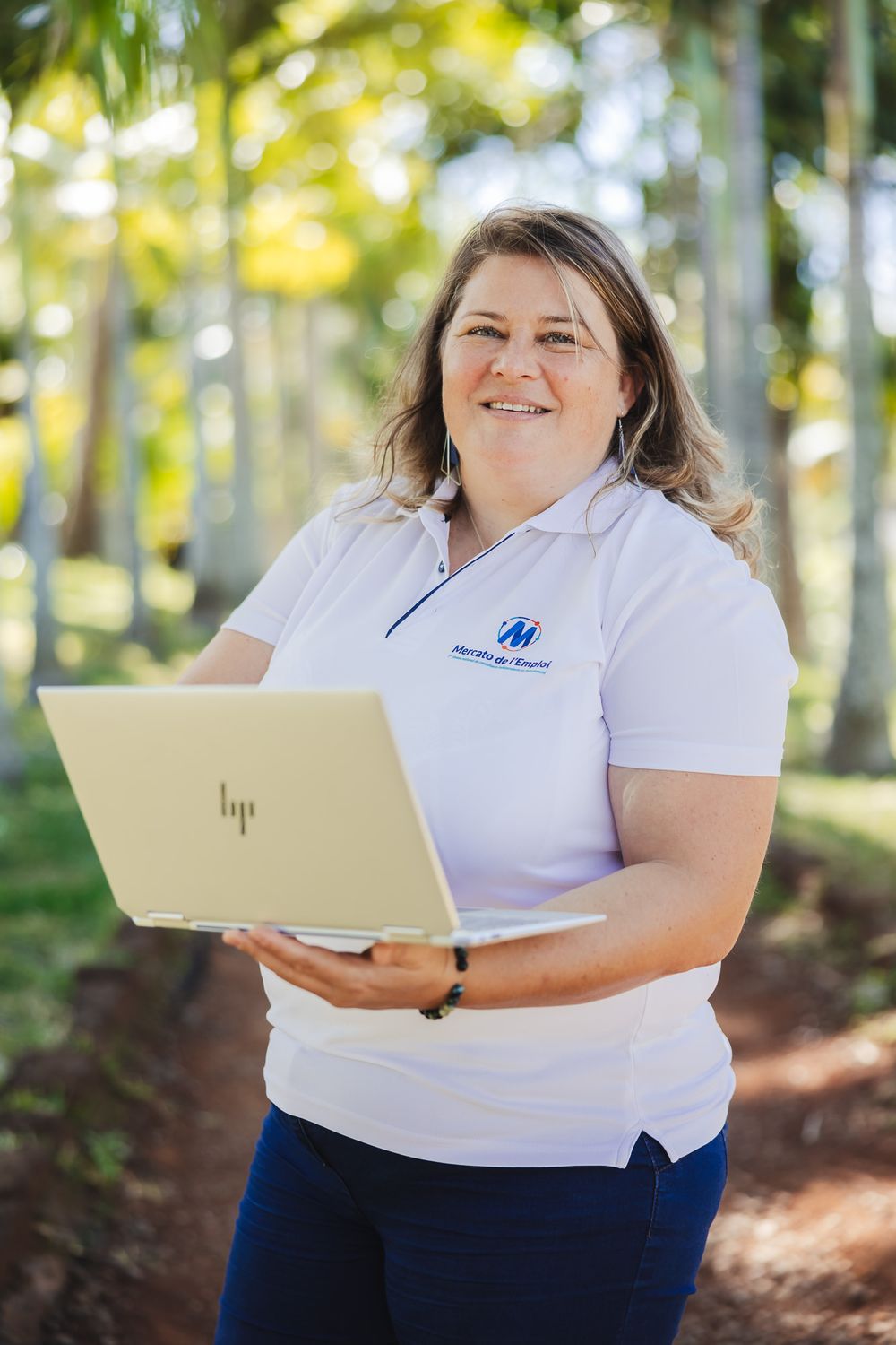 Person in white polo shirt working with laptop outdoors among palm trees.