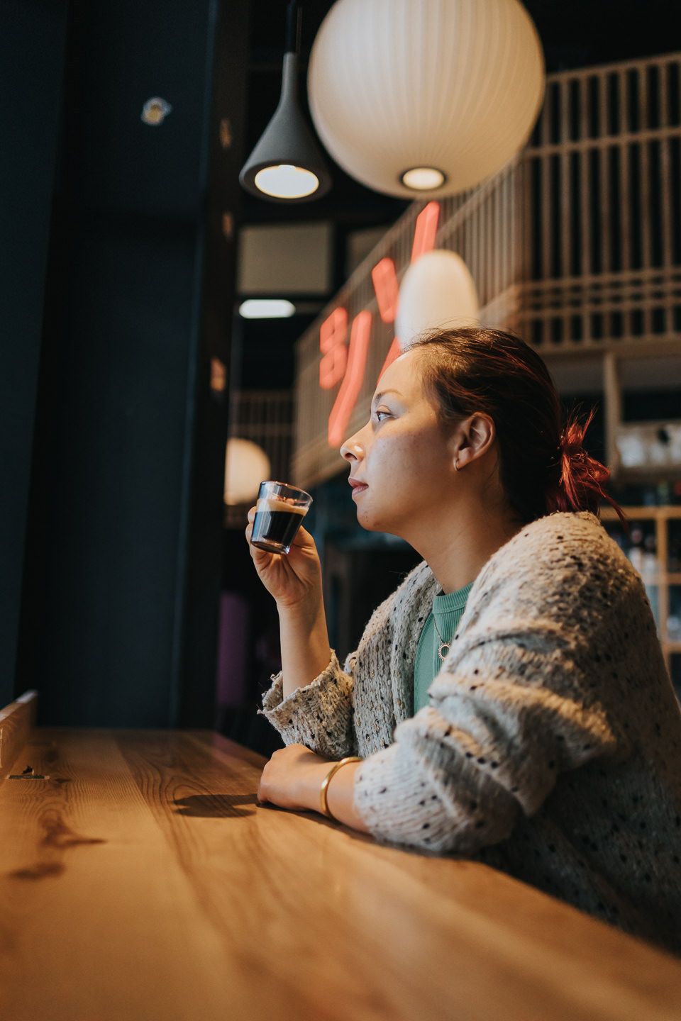 Person in a cozy sweater sitting at a wooden table under pendant lighting in a restaurant.