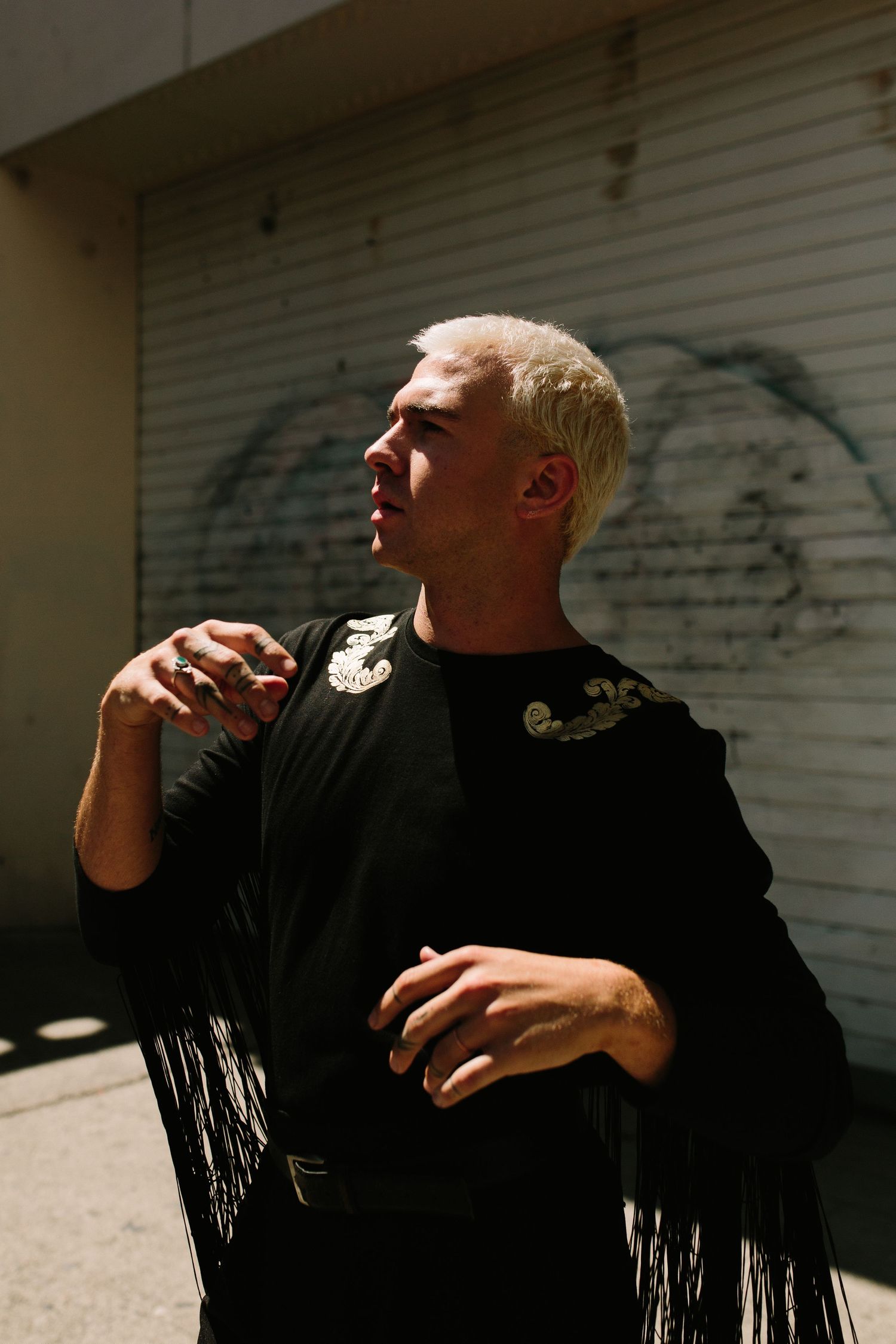 A platinum-haired man in a black fringed jacket poses thoughtfully on a sunny urban street near a graffiti wall.