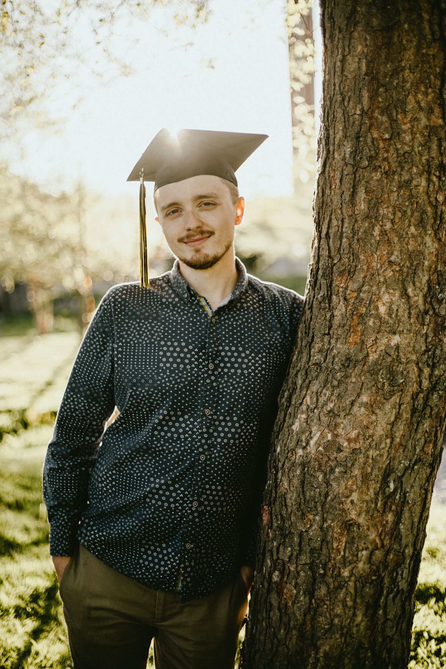 Graduate in patterned shirt poses by tree trunk wearing mortarboard cap in sunlit setting.