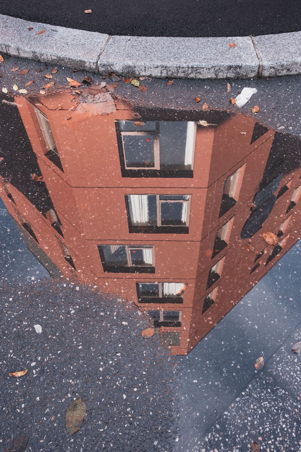 Reflection of a terracotta apartment building in a puddle on wet pavement.