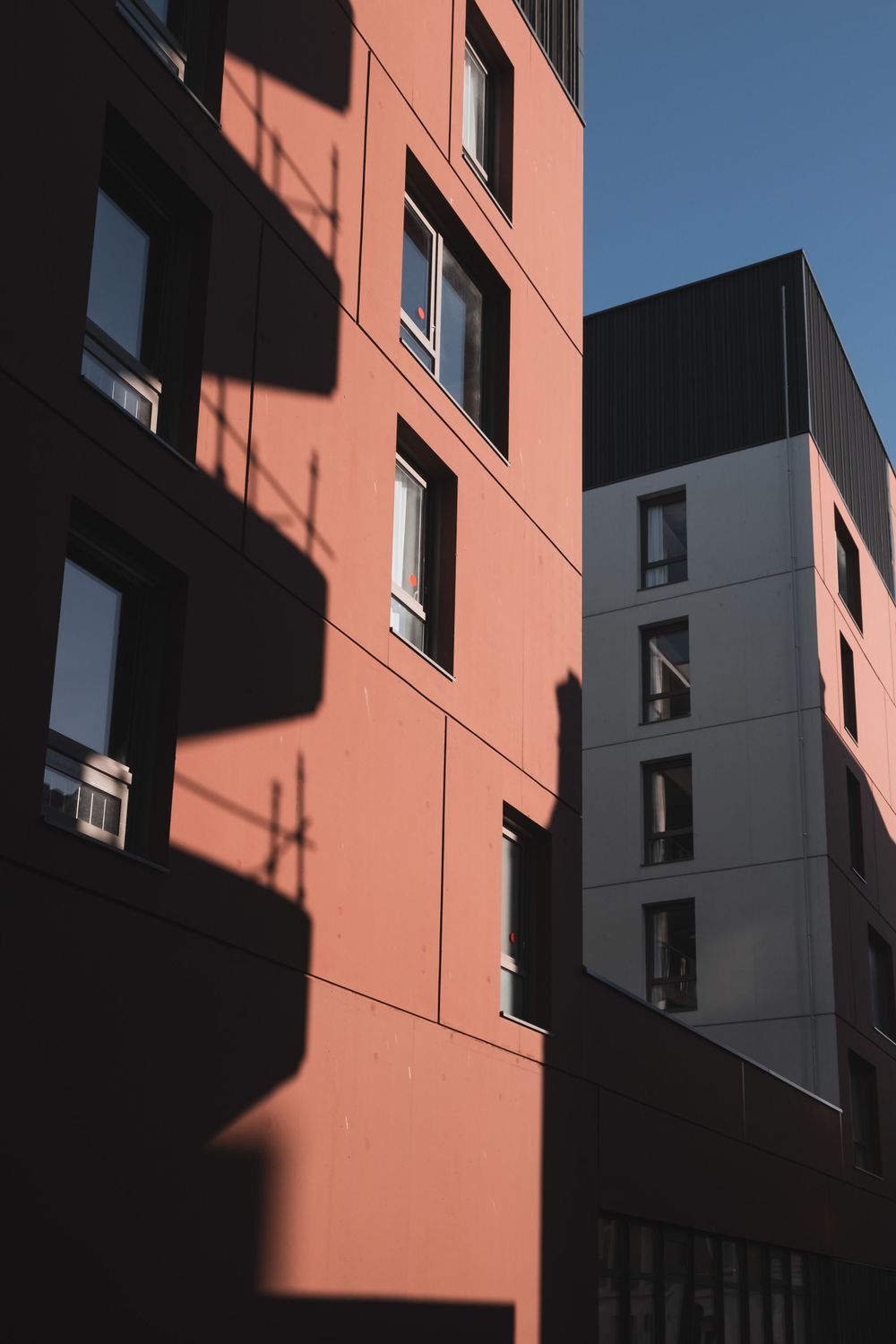Modern apartment building exterior with coral-colored walls and dramatic diagonal shadows.