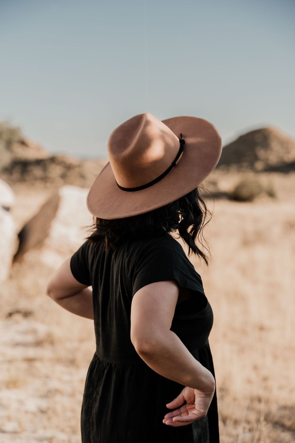 Side profile of person in black dress and brown hat against desert backdrop.