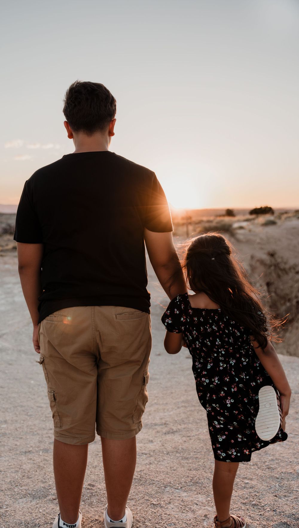 Adult and child walking together at sunset on desert path with sun rays illuminating their silhouettes.