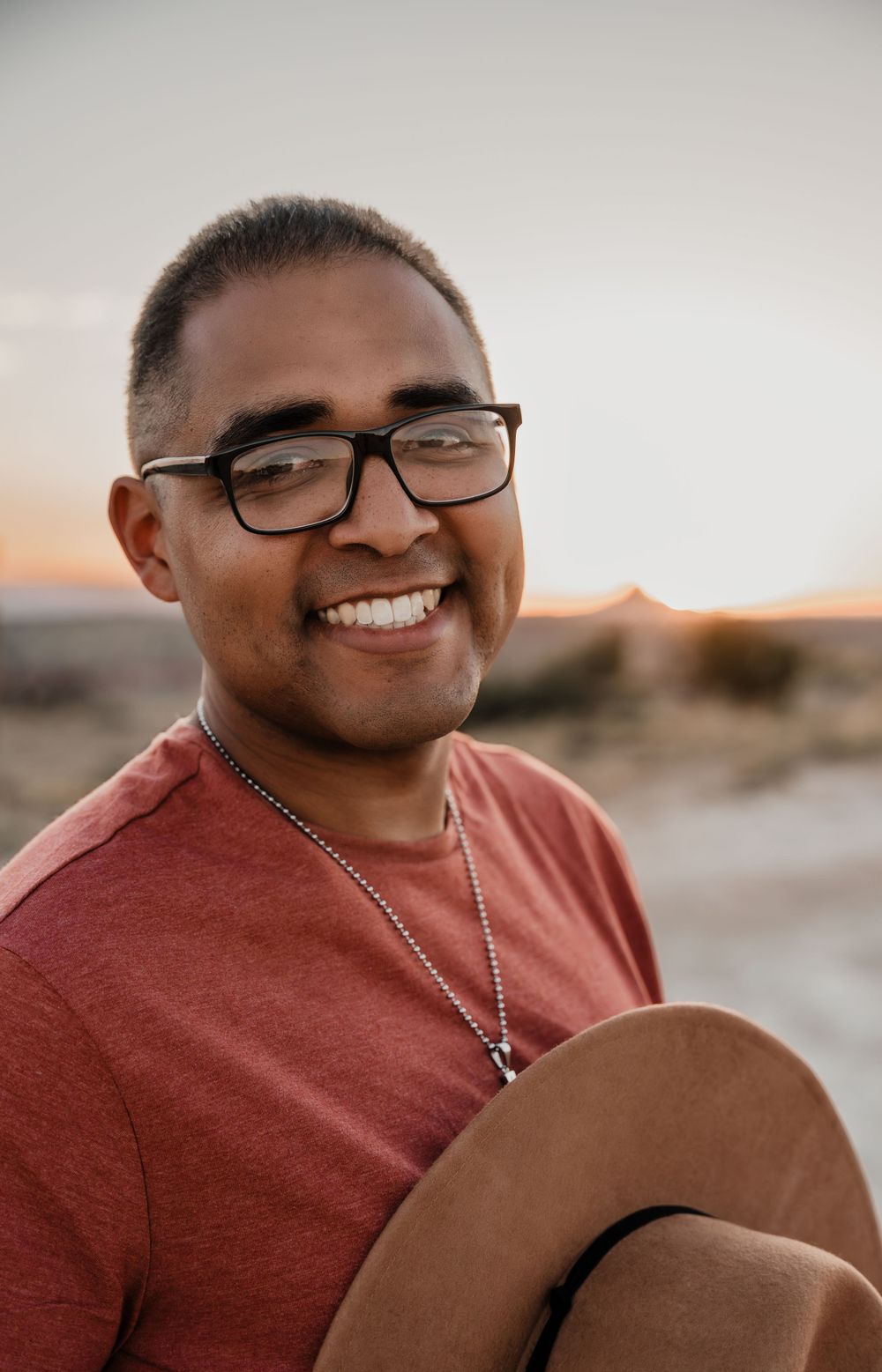 Person wearing glasses and coral shirt smiling warmly during golden hour in desert landscape.