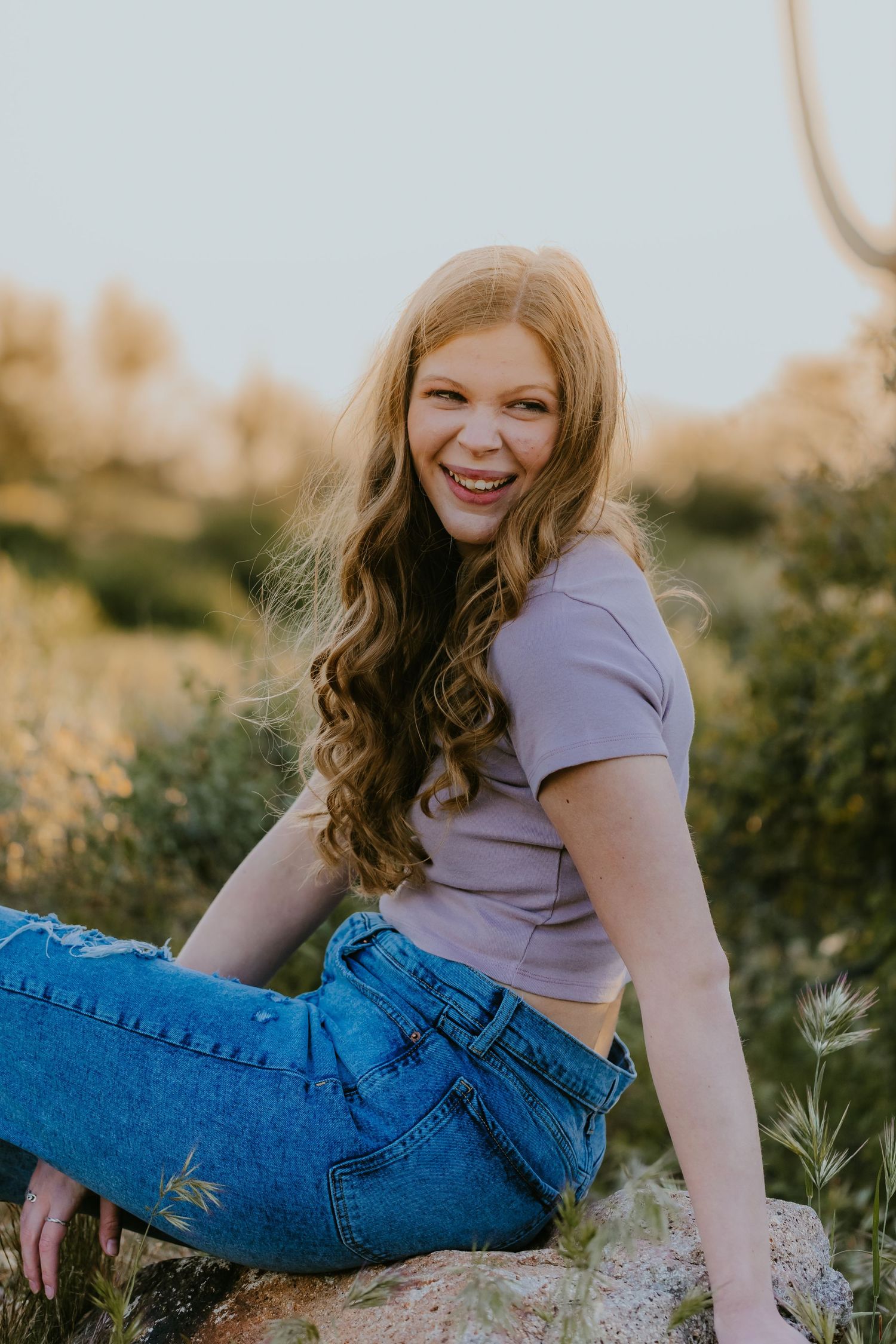 Person in purple top and jeans posing on rock during golden hour photography session.