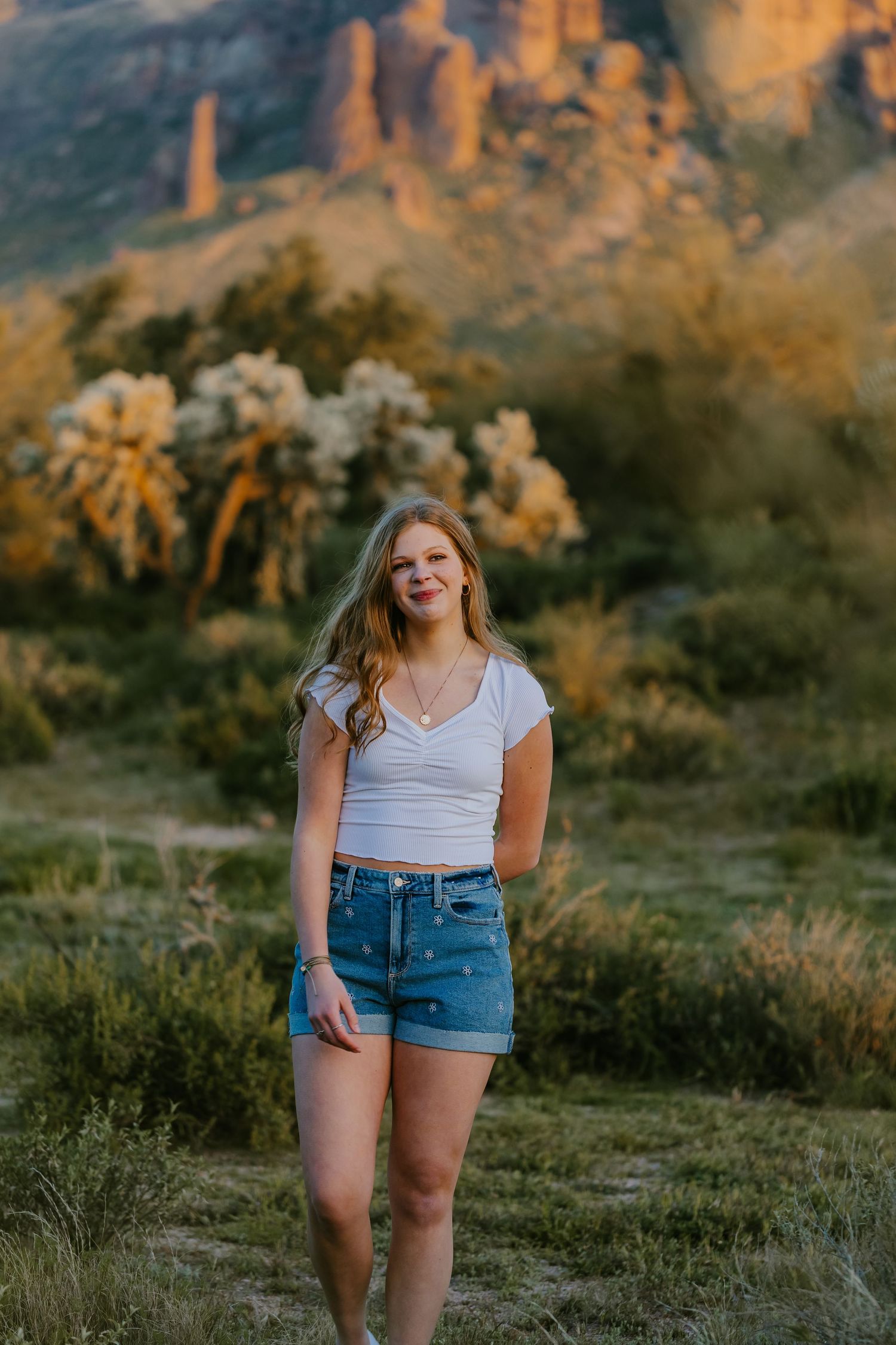 Woman in denim shorts poses in desert landscape with mountains and saguaro cacti at golden hour.